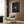 May include: Navy blue poster with gold vinyl record design and spiral text lyrics, displayed in light wood frame on wood paneled wall above cream upholstered chair with geometric pillow, brass floor lamp and dark round side table with open book nearby
