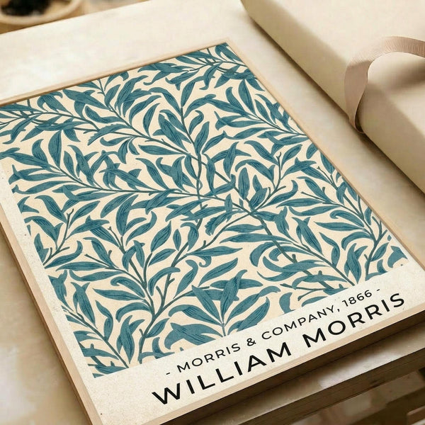 Vintage botanical poster print featuring intricate teal and cream willow branch pattern with flowing leaves and organic curves. The design displays William Morris & Company 1866 text at bottom right corner. Print appears on cream paper with visible texture, photographed alongside neutral packaging materials and cream ribbon on wooden surface.