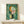 May include: Abstract geometric artwork with green watercolor background featuring circular and rectangular shapes in orange, white, pink, yellow and black. The framed print sits on a light wooden surface next to a small white wrapped gift with natural twine and dried sprig. Modern minimalist room setting with cream walls and natural lighting.