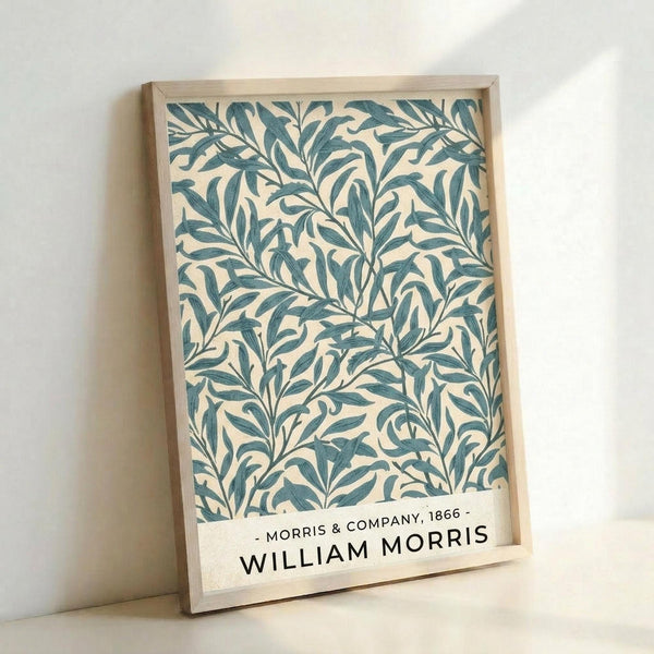 A framed botanical print featuring an intricate pattern of flowing willow branches and leaves in teal blue-green against a cream background. The design shows organic, curving stems with elongated leaves creating an all-over decorative motif. Text at the bottom reads "Morris & Company, 1866 - William Morris" in black lettering. The artwork is displayed in a light wood frame and photographed leaning against a white wall.