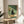 May include: Framed art print displayed on round wooden side table in bright living room with beige chair and plant. Artwork shows black cat with white eyes sitting behind open pizza box containing several slices, with flower vase on green checkered floor. Print has warm green and cream tones in thin natural wood frame.