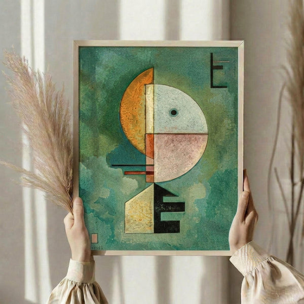 May include: Person holding framed abstract geometric artwork with green textured background, featuring circular and rectangular shapes in orange, white, pink, yellow and black with small dark letters. Light wood frame, hands in cream sweater sleeves visible, dried wheat stems on left side, soft window lighting creates gentle shadows on neutral wall.