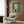 May include: Abstract geometric art print with green background featuring circular and angular shapes in orange, white, pink, and yellow tones, displayed in light wood frame on dark wood paneled wall above cream upholstered armchair with patterned pillow, brass floor lamp and side table with open book nearby