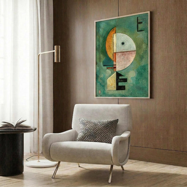 May include: Abstract geometric art print with green background featuring circular and angular shapes in orange, white, pink, and yellow tones, displayed in light wood frame on dark wood paneled wall above cream upholstered armchair with patterned pillow, brass floor lamp and side table with open book nearby