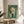 May include: Abstract geometric artwork with green textured background, featuring circular and angular shapes in orange, white, pink, and black with letter "E" visible, displayed in thin wooden frame on round wooden side table next to beige upholstered chair with white pillow, plant leaves visible in corner, natural lighting creating soft shadows on neutral wall.