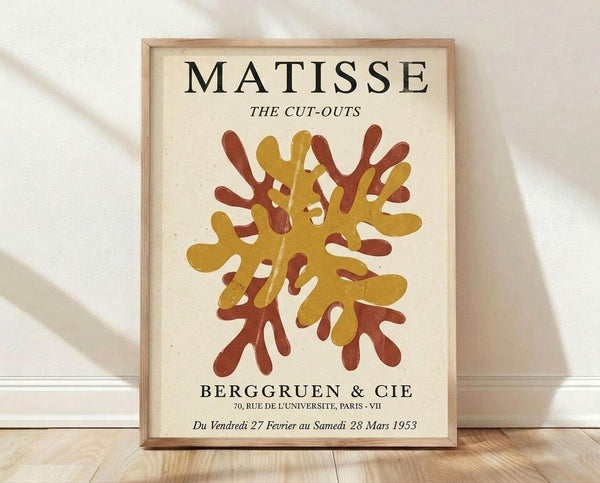 May include: Framed poster with cream background featuring abstract cut-out design in golden yellow and rust red organic shapes resembling coral or seaweed. Black text reads "MATISSE THE CUT-OUTS" at top and "BERGGRUEN & CIE" with Paris gallery information and 1953 exhibition dates below. Light wood frame sits on wooden floor against white wall with soft natural lighting and fabric shadows.