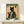 May include: Framed vintage-style poster featuring a black cat with golden eyes wearing a traditional Japanese kimono with floral patterns, holding a red coffee cup. The artwork has aged parchment background with Japanese characters and reads "More Espresso Less Depresso" in cream lettering on green banners. Natural wood frame sits on white wooden furniture beside a small wrapped gift with dried foliage against cream wall.