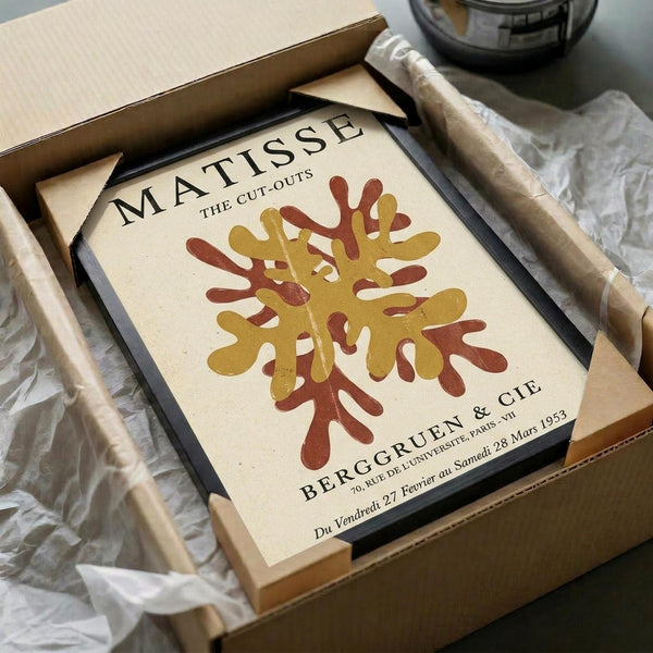 May include: Framed vintage-style art print featuring Matisse's abstract tree design in warm golden yellow and reddish-brown colors on cream background, displayed in black frame inside brown cardboard shipping box with white tissue paper packaging on light surface