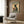 May include: Vintage-style poster featuring a black cat with yellow eyes wearing a traditional Japanese kimono with floral patterns, holding a red tea cup. Text reads "More Espresso Less Depresso" with Japanese characters. Poster is framed and displayed on wood-paneled wall above a modern cream-colored armchair with geometric patterned pillow. Room includes gold floor lamp and dark round side table with open book.