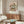 May include: Van Gogh roses poster print in thin gold frame hanging above wooden mid-century credenza in modern living room with beige walls. White curved chair and glass-top coffee table with abstract blue and yellow artwork visible. Poster shows white and pink roses in vase with diagonal pink background pattern.