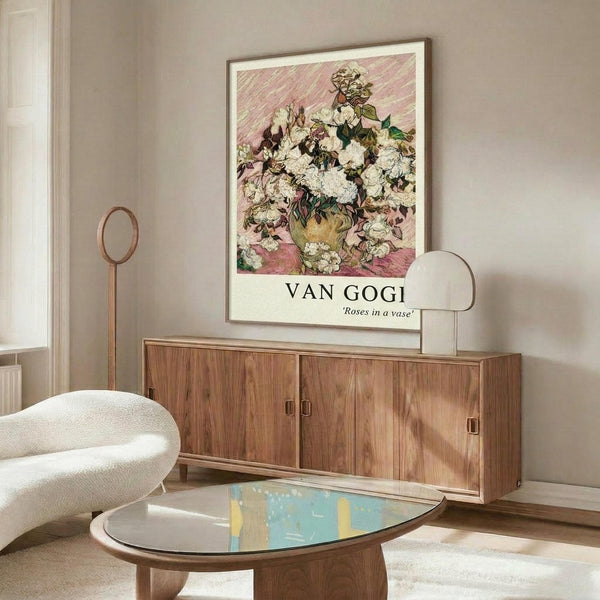 May include: Van Gogh roses poster print in thin gold frame hanging above wooden mid-century credenza in modern living room with beige walls. White curved chair and glass-top coffee table with abstract blue and yellow artwork visible. Poster shows white and pink roses in vase with diagonal pink background pattern.