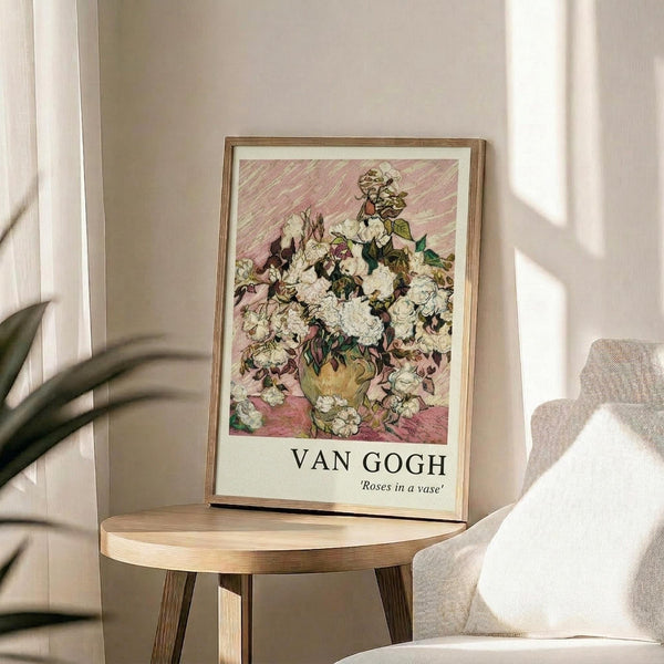 May include: Framed Van Gogh roses poster displayed on round wooden side table next to beige upholstered chair. Print shows white and pink roses in yellow vase against striped pink background, with "VAN GOGH" and "Roses in a vase" text below artwork. Natural lighting creates soft shadows on neutral wall with plant visible in corner.