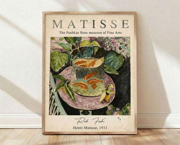 May include: Framed art print featuring Henri Matisse's colorful still life painting with red fish in a glass fishbowl surrounded by lush green foliage and purple flowers. The poster has cream background with "MATISSE" header text and "The Pushkin State museum of Fine Arts" subtitle. Natural wood frame sits on light wooden floor against white wall with soft shadows from window light.