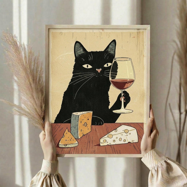May include: Person holding framed poster print showing black cat with white eyes sitting at wooden table, holding wine glass with red wine, surrounded by various cheese wedges. Artwork has vintage cream background with light wooden frame. Dried pampas grass visible on left side of scene.