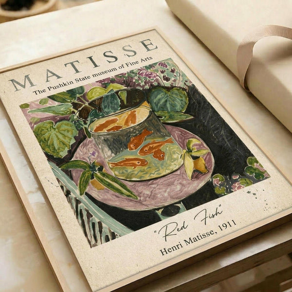 May include: Matisse art print featuring a glass fishbowl with orange goldfish surrounded by lush green foliage and purple flowers on a pink surface. The vintage-style poster shows "MATISSE" and "The Pushkin State Museum of Fine Arts" at the top, with "Red Fish Henri Matisse, 1911" at the bottom. The print sits on cream-colored paper with neutral ribbon visible, creating an elegant flat lay presentation.