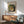 May include: Framed vintage art poster featuring a colorful still life painting with goldfish in a glass bowl surrounded by green foliage, displayed above a mid-century modern wooden sideboard with sliding doors. Scene includes a sage green upholstered armchair with wooden arms, white ceramic table lamp, and round coffee table. Natural sunlight creates geometric shadows on white wall.