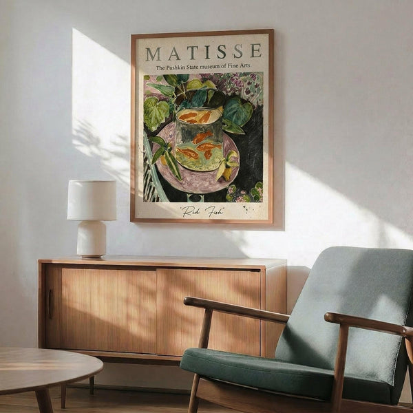 May include: Framed vintage art poster featuring a colorful still life painting with goldfish in a glass bowl surrounded by green foliage, displayed above a mid-century modern wooden sideboard with sliding doors. Scene includes a sage green upholstered armchair with wooden arms, white ceramic table lamp, and round coffee table. Natural sunlight creates geometric shadows on white wall.