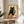 May include: Framed poster print displayed on round wooden side table next to beige upholstered chair and plant. Artwork shows black cat with green eyes holding wine glass, surrounded by cheese pieces on wooden surface. Vintage-style illustration with cream background in natural wood frame. Soft natural lighting creates shadows across neutral-toned room setting.