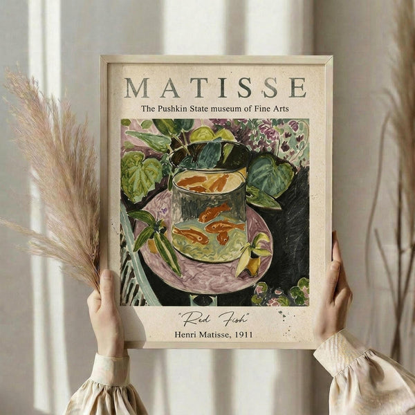 May include: Person in cream outfit holding framed art print against white curtained window, with dried wheat stems. Print shows Matisse's colorful still life painting of red goldfish in glass bowl surrounded by lush green plants on purple surface. Light wooden frame with museum exhibition styling and artwork details below image.