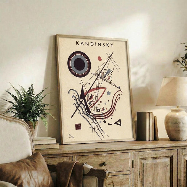May include: Framed abstract art print with geometric shapes, circles, and lines in black, red, and burgundy on cream background, displayed on rustic wooden console with books, ceramic vase, and fern plant in modern living room setting with beige upholstered chair