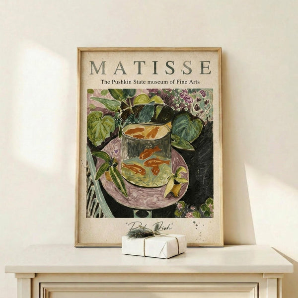May include: Framed vintage art poster featuring a colorful still life painting of goldfish in a glass bowl surrounded by lush green plants and purple flowers. The cream-colored poster displays "MATISSE" and "The Pushkin State museum of Fine Arts" text above the artwork. The wooden frame sits on a light wood shelf against a white wall, with a small wrapped gift box beside it.
