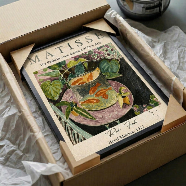 May include: Framed art print featuring Henri Matisse's colorful still life painting with goldfish in a glass bowl, surrounded by lush green plants and purple flowers. The vintage museum poster shows vibrant oranges, greens, and purples against a dark background. The black-framed print sits in an open cardboard shipping box with white tissue paper, alongside wooden packing materials on a light surface.