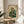 May include: Framed poster print displayed on wooden round side table next to beige upholstered chair in sunlit room with white walls and plant shadows. Artwork shows anthropomorphic green frog wearing ornate teal and gold patterned kimono, smoking a pipe with swirling smoke, surrounded by bamboo leaves. Japanese characters visible in upper right corner. Warm natural lighting creates soft shadows across the scene.