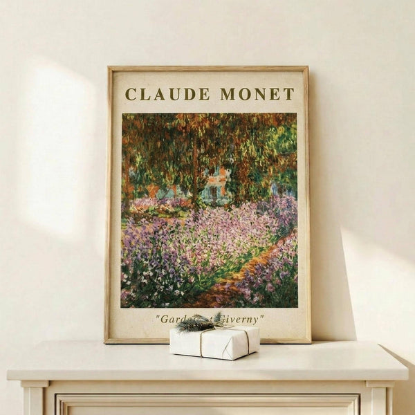 May include: Framed poster print of Claude Monet's impressionist garden painting featuring vibrant purple irises in foreground, lush green foliage and trees overhead, displayed on light wooden console table beside small white wrapped gift with evergreen sprig against cream wall