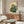 May include: Vintage-style Japanese woodblock print featuring a green frog in traditional blue and gold kimono smoking a long pipe, displayed in light wooden frame above mid-century walnut credenza with sliding doors. Modern white curved chair and glass-top oval coffee table complete the minimalist room setting with white walls and natural lighting.