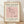 May include: Framed poster print leaning against white textured wall on light wood floor. Wood frame contains cream-colored print with pink square featuring minimalist black line drawing of flamingo. Text reads "PICASSO LINE DRAWINGS & PRINTS" at top and "LE FLAMAND ROSE Les Métamorphoses, 1930" at bottom. Soft natural lighting creates gentle shadows on wall.