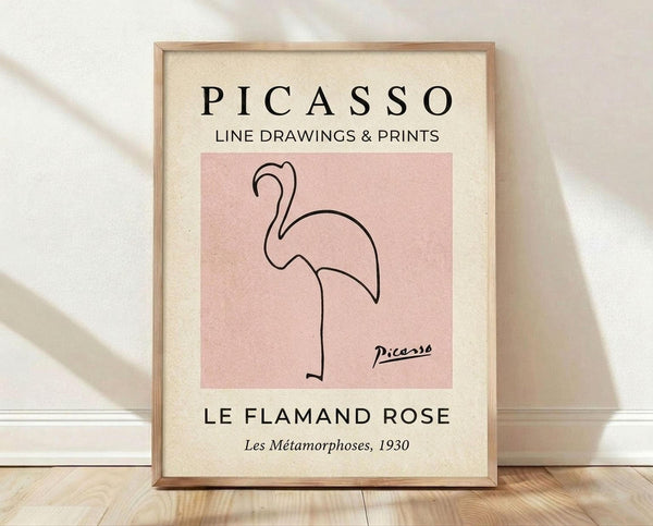 May include: Framed poster print leaning against white textured wall on light wood floor. Wood frame contains cream-colored print with pink square featuring minimalist black line drawing of flamingo. Text reads "PICASSO LINE DRAWINGS & PRINTS" at top and "LE FLAMAND ROSE Les Métamorphoses, 1930" at bottom. Soft natural lighting creates gentle shadows on wall.