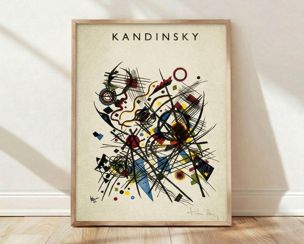 May include: Framed abstract art print in natural wood frame leaning against white wall on light wooden floor. The artwork features dynamic black lines, geometric shapes, and colorful elements in red, blue, yellow, and green on cream background. Artist name "Kandinsky" appears at top with signature at bottom. Soft natural lighting creates subtle shadows on the wall.