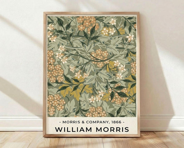 May include: Framed botanical art print with William Morris design featuring sage green background with intricate floral pattern in coral, cream and gold tones. Delicate branches, leaves and small clustered flowers create an ornate vintage wallpaper style. Light wood frame sits on wooden floor against white textured wall with soft natural lighting casting shadows.