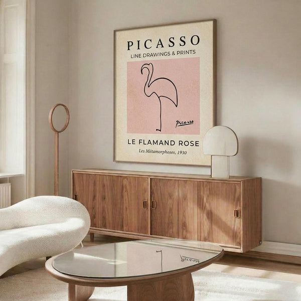 May include: Modern living room with walnut sideboard displaying framed Picasso flamingo line drawing poster. Pink minimalist artwork shows single-line flamingo illustration on cream background in thin wooden frame. Scene features white curved chair, glass coffee table, brass floor lamp, and neutral wall colors with natural lighting.