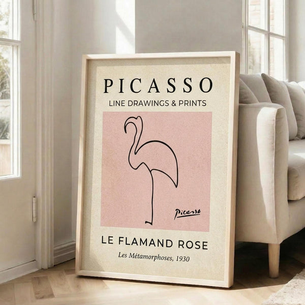 May include: Framed art print leaning against white wall next to beige sofa in bright living room with large windows. Print features minimalist line drawing of pink flamingo on dusty rose background with cream border, displayed in light wood frame on hardwood floor.
