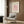 May include: A framed art print displayed on wood paneling above a beige upholstered chair. The poster features a minimalist line drawing of a flamingo in black on a soft pink background, with cream-colored borders and text identifying it as a Picasso work. A brass floor lamp and dark round side table with an open book complete the modern interior scene.