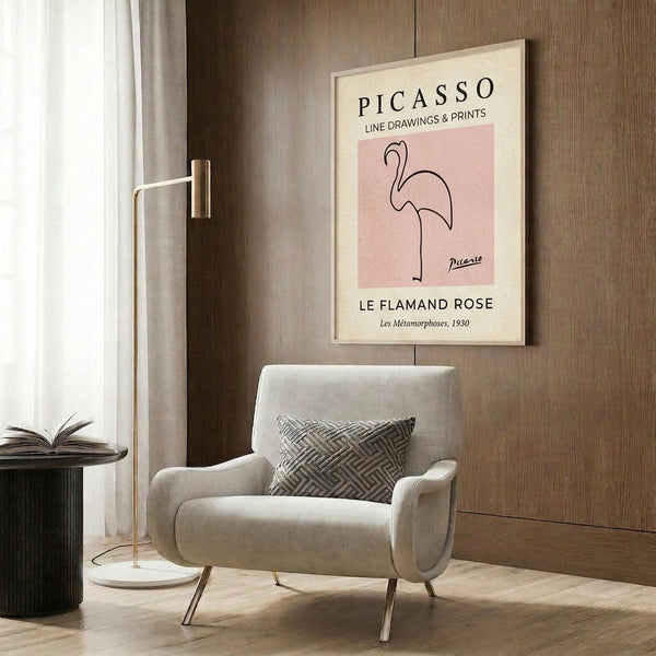May include: A framed art print displayed on wood paneling above a beige upholstered chair. The poster features a minimalist line drawing of a flamingo in black on a soft pink background, with cream-colored borders and text identifying it as a Picasso work. A brass floor lamp and dark round side table with an open book complete the modern interior scene.