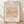 May include: A framed art print leaning against a white textured wall on light wooden flooring. The poster features Picasso's minimalist line drawing of a dachshund dog in black ink on a peachy-pink background. Text reads "PICASSO LINE DRAWINGS & PRINTS" at top and "LE CHIEN Les Métamorphoses, 1930" at bottom on cream-colored paper. The simple wooden frame has natural wood tones. Soft shadows fall across the scene.