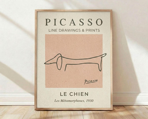 May include: A framed art print leaning against a white textured wall on light wooden flooring. The poster features Picasso's minimalist line drawing of a dachshund dog in black ink on a peachy-pink background. Text reads "PICASSO LINE DRAWINGS & PRINTS" at top and "LE CHIEN Les Métamorphoses, 1930" at bottom on cream-colored paper. The simple wooden frame has natural wood tones. Soft shadows fall across the scene.