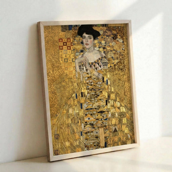 May include: Framed reproduction of Gustav Klimt's golden portrait painting showing an elegant woman in ornate dress against decorative patterned background. The artwork features rich gold tones, geometric and spiral motifs, and Art Nouveau styling. Simple light wood frame displayed leaning against white wall on white surface, showing the print's warm metallic colors and intricate details.