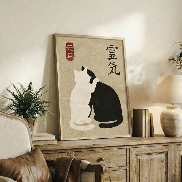 May include: Framed poster with beige background featuring black and white cats in yin yang formation, red and black Japanese characters above. Wooden frame rests on rustic wood sideboard beside fern plant, books, ceramic vases. Cream chair with brown throw blanket visible in cozy living room setting with white walls.