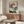 May include: Japanese woodblock print artwork featuring white cranes by water with pine tree branches and orange sun, displayed in thin wooden frame above mid-century walnut credenza. Modern living room setting with cream textured chair, glass-top oval coffee table, and minimalist brass floor lamp on light walls.