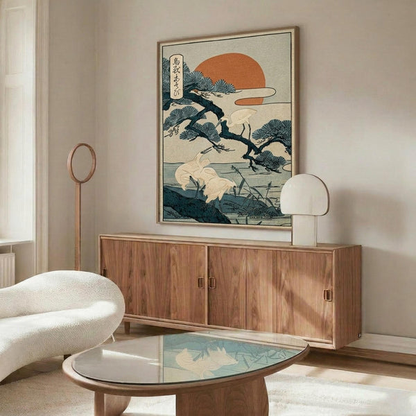 May include: Japanese woodblock print artwork featuring white cranes by water with pine tree branches and orange sun, displayed in thin wooden frame above mid-century walnut credenza. Modern living room setting with cream textured chair, glass-top oval coffee table, and minimalist brass floor lamp on light walls.