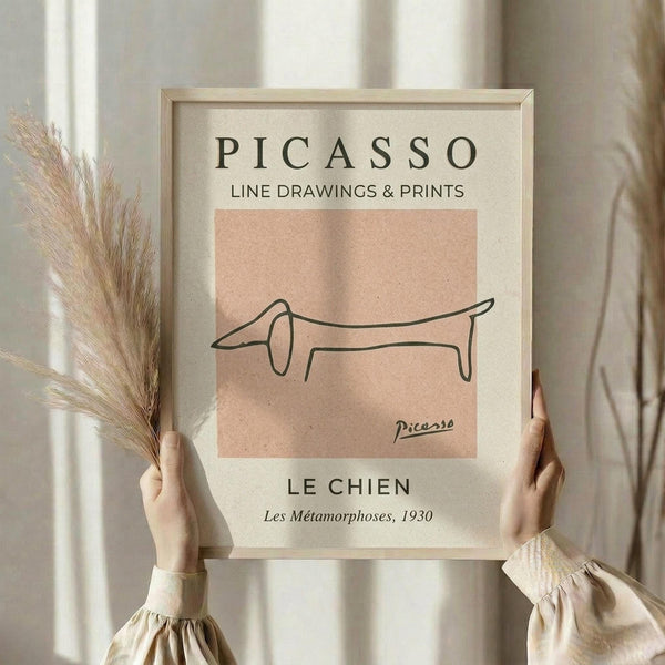May include: Hands holding a framed art print featuring Picasso's minimalist line drawing of a dachshund dog in black lines on a peach background. The cream-colored print includes exhibition text above and below the artwork. Person wears a beige ruffled sleeve top and holds dried pampas grass, photographed against soft neutral curtains in natural lighting.