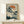 May include: Japanese woodblock print artwork in light wood frame showing white cranes near pine tree with orange sun and blue-green foliage, displayed on light wood console table with small white wrapped gift, against cream wall in minimalist room setting.