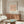 May include: Minimalist line drawing of a dachshund dog on peach background with white text reading "PICASSO LINE DRAWINGS & PRINTS" and "LE CHIEN Les Métamorphoses, 1930" displayed in thin wooden frame above walnut credenza. Modern living room setting with cream sculptural chair, glass coffee table with colorful abstract art underneath, white table lamp, and brass mirror on neutral walls.