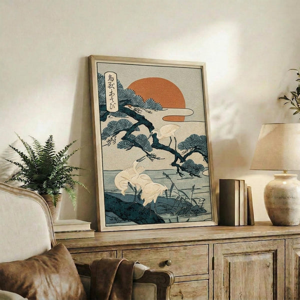 May include: Japanese woodblock print featuring white cranes near water with pine tree branches, orange sun, and Japanese text, displayed in light wood frame on rustic wooden sideboard alongside fern plant, ceramic vase, and books in neutral-toned living room setting.