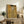 May include: Gold-toned reproduction of Klimt's portrait showing a woman in ornate patterned dress against decorative background, displayed in light wooden frame on rustic wooden sideboard. Room features neutral walls, fern plant, ceramic vase, books, and brown leather chair. Artwork shows characteristic Art Nouveau style with geometric patterns and golden hues.