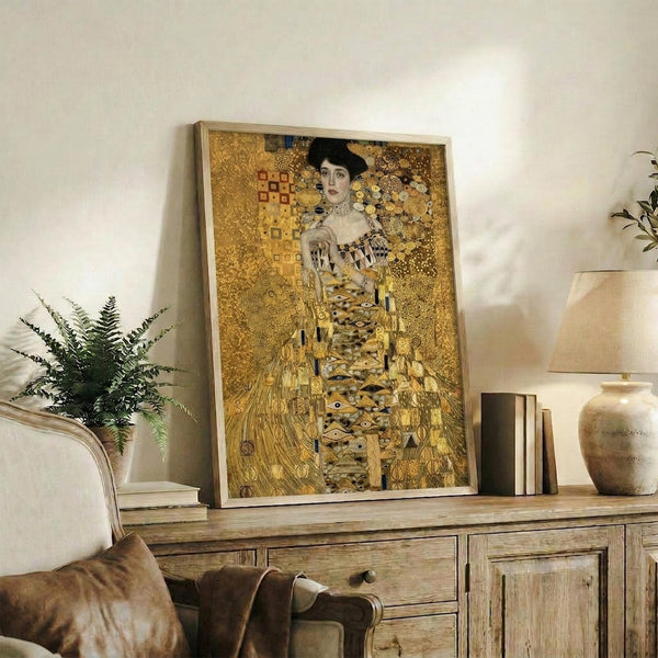 May include: Gold-toned reproduction of Klimt's portrait showing a woman in ornate patterned dress against decorative background, displayed in light wooden frame on rustic wooden sideboard. Room features neutral walls, fern plant, ceramic vase, books, and brown leather chair. Artwork shows characteristic Art Nouveau style with geometric patterns and golden hues.