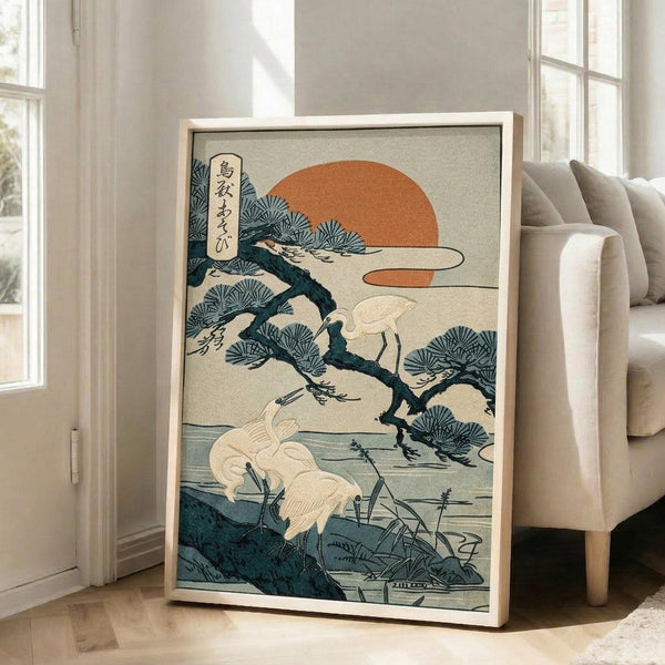 May include: A framed Japanese woodblock print featuring white cranes perched on pine branches with an orange sun in the background, displayed on a wooden floor next to a beige upholstered chair in a bright room with large windows. The artwork shows traditional ukiyo-e style with teal pine needles, cream-colored birds wading in water below, and Japanese text in the upper left corner, all set within a light wooden frame.